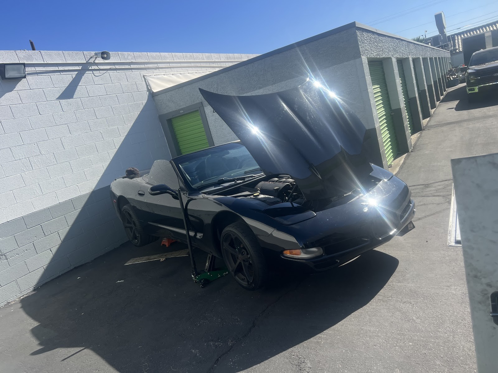 Black Corvette convertible with hood up on a jack outdoors near storage units