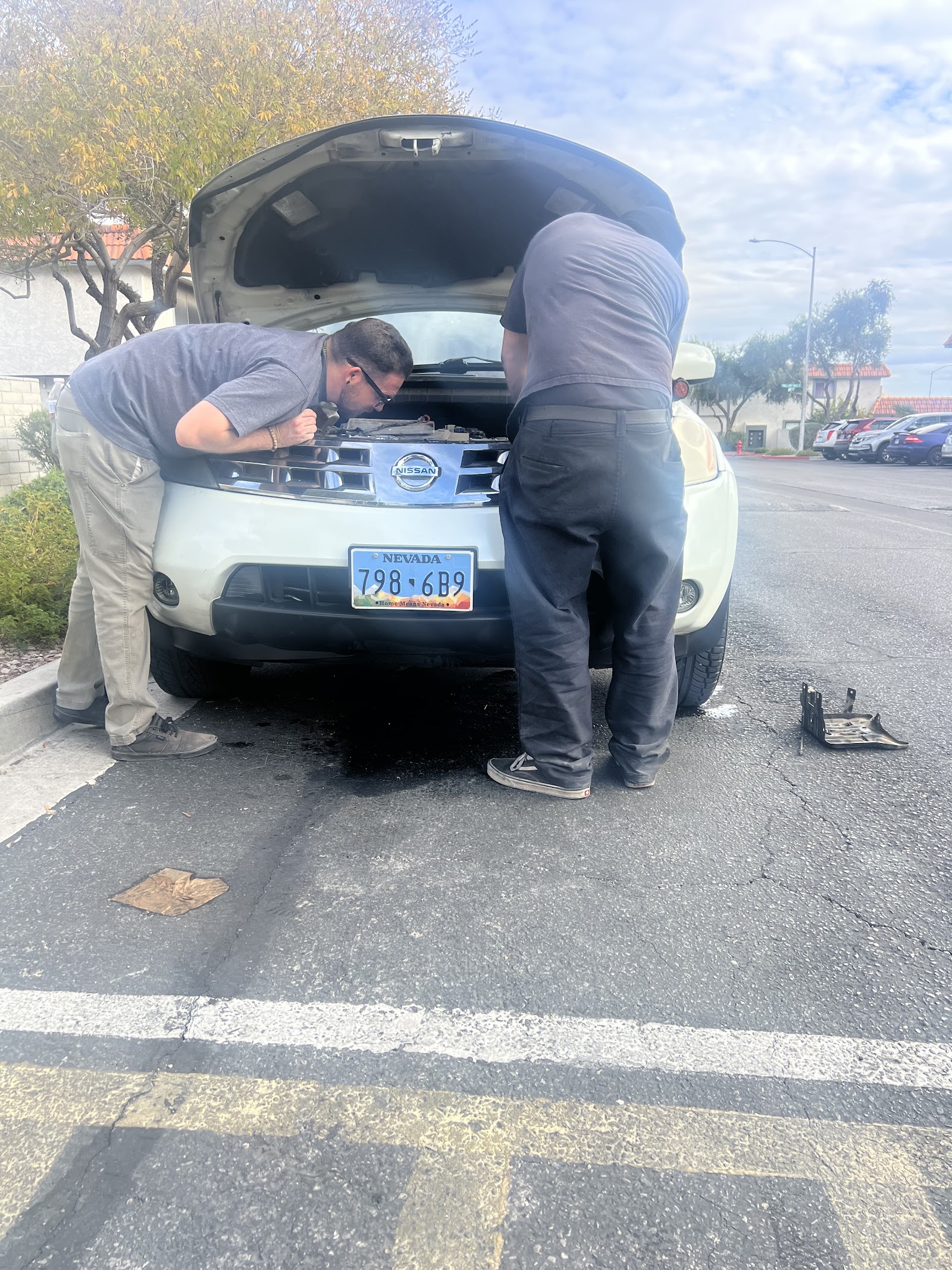 Two mechanics working under the hood of a white Nissan SUV in a parking lot